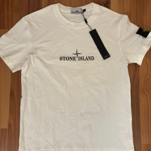 Stone Island t-shirt - Vit t-shirt från Stone Island i bomull med klassisk logga tryckt på bröstet och stor kompassgrafik på ryggen. Ikonisk patch med broderad logga på ärmen. Enkel och clean design, perfekt för dig som gillar streetwear.