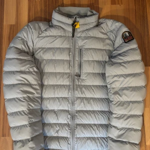 Parajumpers jacka - Ljusgrå dunjacka från Parajumpers i superlätt material. Jackan har quiltad design, hög krage, dragkedja framtill och en bröstficka med dragkedja. Klassisk PJS-logga på ärmen och cool gul detalj vid halsen. Perfekt för kalla dagar när du vill hålla stilen.