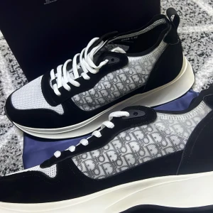 Dior B25 svart vita - Snygga Dior sneakers med svart och vit färg, mesh och skinnmaterial samt tydligt Dior-monogram i grått på sidorna. Skorna har rund tå, platt sula och vita snören. Perfekt för dig som gillar lyxiga detaljer och modern design.