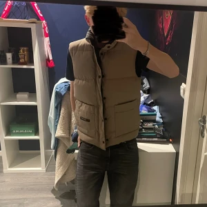 En beige Vintage dunväst från Canada Goose - En beige dunväst köpt 2003. En väldigt varm väst som passar perfekt till hösten och vintern. Storleken är S och han på bilden är 178 cm lång. Vesten går för runt 1500 kronor på vestaire collection. Den är nästintill oanvänd och har bara suttit i garderoben. 