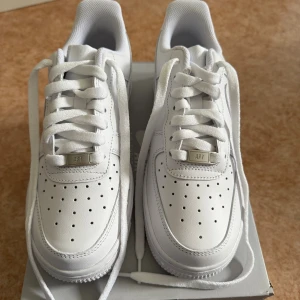 Nike Air Force 1 vita sneakers - Nike Air Force 1 sneakers i helvitt i storlek 38 med klassisk design och perforerad tå. Skorna har snörning, platt sula och rund tå. Skorna har aldrig använts och har kvar originalförpackningen. 