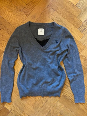 Grå v-ringad tröja Abercrombie & Fitch - Mjuk och stilren grå stickad tröja från Abercrombie & Fitch med v-ringning och diskret mörkblå logga broderad på bröstet. Tröjan har långa ärmar och ribbade muddar vid ärmslut och nederkant. Perfekt för lager på lager och enkel att matcha.