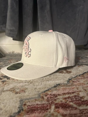 Vit New Era White Sox keps - Snygg vit keps från New Era med broderad White Sox-logga i ljusrosa framtill och en unik patch med blommor på sidan. Kepsen har rak skärm och klassisk snapback-modell. Perfekt för dig som vill sticka ut med stilrena detaljer.