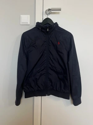 Vindjacka från Polo Ralph Lauren - Marinblå vindjacka från Polo Ralph Lauren i nyskick. Jackan är i storlek 14-16 år, och har en luva som går att gömma. Perfekt för blåsiga dagar samtidigt som den passar med alla typer av outfits.