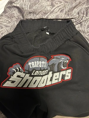 Trapstar London Shooters hoodie svart - Svart hoodie från Trapstar med stort broderat motiv på bröstet där det står 'Trapstar London Shooters' och en panter. Hoodien har huva och ribbade muddar. Materialet är mjukt och känns som bomullsblandning. Perfekt för dig som gillar streetwear och vill sticka ut.