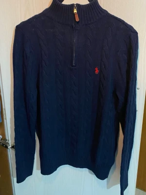 Mörkblå kabelstickad tröja Polo Ralph Lauren - Mörkblå kabelstickad tröja från Polo Ralph Lauren med halv dragkedja och hög krage. Tröjan har det klassiska röda broderade logotypen på bröstet och ribbstickade muddar. Perfekt för en stilren och avslappnad look.