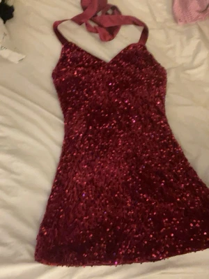 Vinröd glittrig kort klänning - Säljer en vinröd kort klänning med paljetter och halterneck-band. Klänningen har en v-ringad urringning och är täckt av glittriga paljetter som ger en festlig look. Perfekt för dig som vill sticka ut med en snygg och glammig stil.