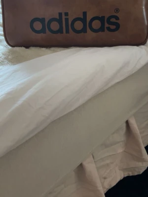 Retro brun resväska från Adidas - Snygg retro resväska från Adidas i brunt konstläder med stor svart logga på framsidan. Väskan har en klassisk rektangulär form och metallfötter undertill för extra stabilitet. Perfekt för dig som vill resa med stil och gillar vintagevibbar.