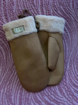 Beige tumvantar från UGG med päls - Mysiga beige tumvantar från UGG med mjukt foder och fluffig pälsdetalj vid mudden. Utsidan är i mocka och insidan är supergosig, perfekta för kalla dagar. Klassisk design med snöre mellan vantarna.