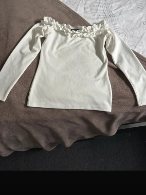 Vit långärmad topp med volang - Söt vit långärmad topp med volangdetalj längs den offshoulder-inspirerade halsringningen. Tillverkad i mjukt material som känns bekvämt mot huden. Perfekt för dig som gillar en feminin och stilren look.