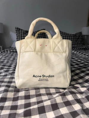 Vit canvasväska Acne Studios - Säljer en stilren vit handväska från Acne Studios i canvas med både handtag och axelrem. Väskan har Acne Studios-logga tryckt framtill. Den är köpt på Vestiaire Collective med äkthetskontroll (se bild) Det finns några fläckar på den men de är mest på handtagen och insidan av väskan. Kan skicka fler bilder på det om behövs! 