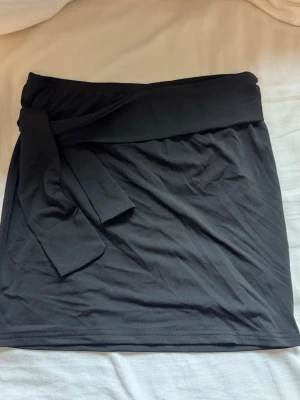 Svart kortkjol med knyt i midjan - Stilren svart kortkjol med bred linning och knytband i midjan. Kjolen har en enkel, rak passform och är tillverkad i ett mjukt material som känns bekvämt mot huden. Perfekt för dig som gillar minimalistisk stil och vill ha en kjol som är lätt att matcha.