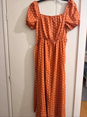 Orange prickig långklänning med puffärm - Superfin orange långklänning med vita prickar och puffärmar. Klänningen har en markerad midja med tillhörande bälte och fyrkantig urringning. Perfekt för sommarens alla tillfällen och ger en härlig retrovibe.