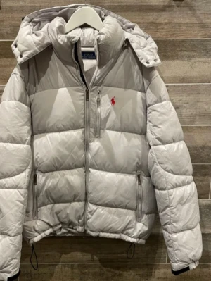 Ljusgrå pufferjacka från Polo Ralph Lauren - Snygg ljusgrå pufferjacka från Polo Ralph Lauren med röd logga på bröstet. Jackan har huva, dragkedja framtill, två sidofickor med dragkedja och quiltad design. Perfekt för kalla dagar och riktigt trendig look.