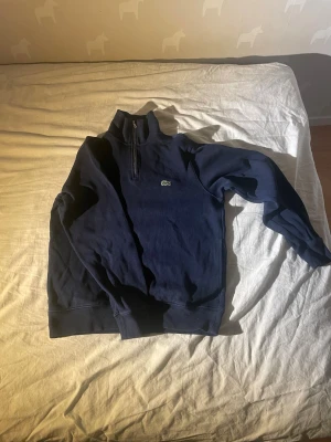 Mörkblå halfzip från Lacoste - En lite sjystare Mörkblå tröja från Lacoste med halv dragkedja och klassisk krokodillogga på bröstet. Tröjan har hög krage, ribbade muddar och är tillverkad i mjukt material. Perfekt för en avslappnad och stilren look. Använd få tal gånger då den är lite liten för mig. 