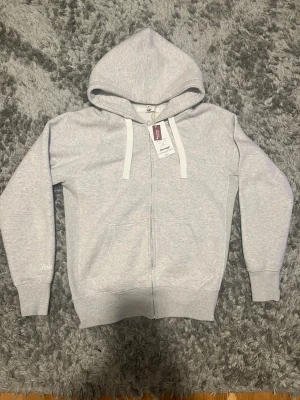 Ljusgrå hoodie från Russemerch - Snygg ljusgrå hoodie från Russell Athletic med dragkedja framtill, två fickor och justerbar huva med vita snören. Tillverkad i mjukt bomullsmaterial som känns skönt mot huden. Perfekt för en avslappnad och stilren look.