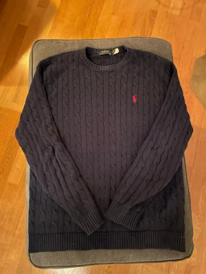 Mörkblå kabelstickad tröja Polo - Mörkblå kabelstickad tröja från Polo Ralph Lauren med rund halsringning och röd broderad logga på bröstet. Tröjan har långa ärmar och ribbade muddar vid ärmslut och nederkant. Färgen är marinblå och den passar M och L. Priset går att diskutera.