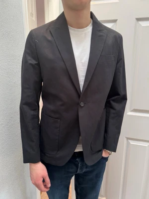 Svart kavaj med enkel knapp - Stilren svart kavaj med klassisk passform och enkelknäppning. Kavajen har två stora framfickor och en bröstficka. Materialet ser ut att vara bomull eller en bomullsblandning. Perfekt för en clean och modern look.