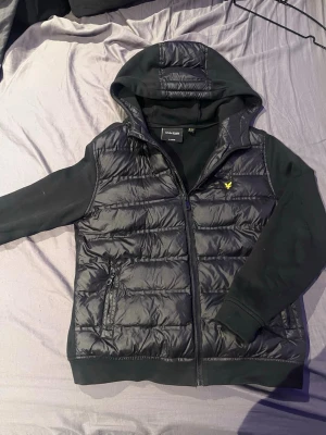 Svart cardigan från Lyle & Scott - Snygg svart cardigan från Lyle & Scott med huva och dragkedja framtill. Västen har quiltad design, två fickor med dragkedja och den klassiska gula loggan på bröstet. Perfekt för lager-på-lager och streetwear-stil.