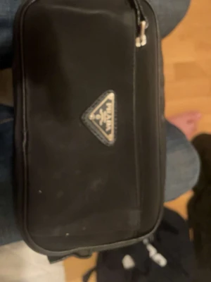 Svart axelväska från Prada - Den är smutsig i bilder men i realise har tvättat den o kmr skicka den ren Snygg svart axelväska från Prada i klassisk rektangulär form. Väskan har silverfärgade detaljer och dragkedja samt det ikoniska Prada-emblemet framtill. Tillverkad i slitstarkt syntetmaterial med justerbar axelrem.