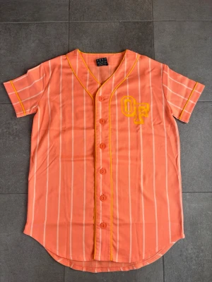 Odd future baseballtröja  - Säljer en orange baseballinspirerad skjorta från OF med vita vertikala ränder och gula detaljer. Skjortan har korta ärmar, knappar framtill och ett gult OF-tryck på bröstet. Perfekt för dig som vill sticka ut med en sportig vibe.