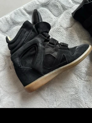 Svarta kilklackade stövletter i mocka - Säljer ett par svarta stövletter med kilklack och kardborreband. Skorna är gjorda i mocka och har en cool, sportig design med gummisula. Perfekta för dig som vill ha en edgy look och gillar streetstyle.