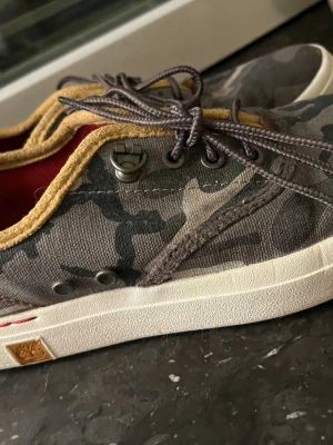 Camouflage  från Timberland - Säljer ett par snygga  skor  från Timberland med grått camouflage-mönster. Skorna har beige kant, röda detaljer på insidan och vita sulor. Tillverkade i slitstark bomull med klassiska snörningar och metallöglor. Perfekta för en avslappnad streetstil.