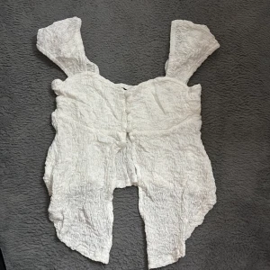 Vit topp med knappar - Söt vit topp med omlottdesign och knappar framtill. Gjord i ett skrynkligt, lätt material med breda axelband och asymmetrisk nederkant. Perfekt för dig som gillar en romantisk och avslappnad stil.