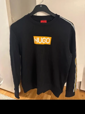 Svart sweatshirt från Hugo Boss M - Svart långärmad sweatshirt från Hugo Boss med gul och vit logga på bröstet. Snygga band med HUGO-tryck i vitt och gult längs ärmarna. Rund hals och normal passform. Materialet är mjuk bomull, perfekt för chill dagar.
