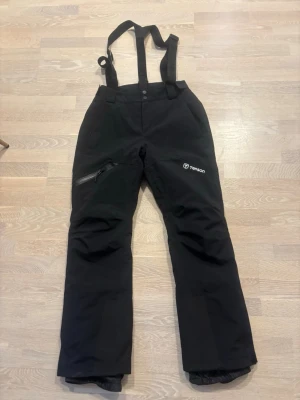 Svarta skidbyxor från Tenson - Säljer ett par svarta täckbyxor från Tenson med hängslen och dragkedja vid benet. Byxorna har två framfickor och en extra ficka med dragkedja på benet. Perfekta för vinterns alla äventyr i snön.
