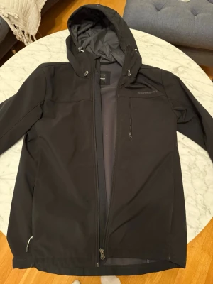 Svart vindjacka Peak Performance - Snygg svart vindjacka från Peak Performance med huva och dragkedja framtill. Jackan har en bröstficka med dragkedja och justerbar huva. Tillverkad i ett slitstarkt, vindtätt material som passar perfekt för blåsiga dagar.