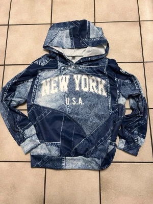 Blå hoodie med New York-tryck - Snygg hoodie med blått denim-mönster och stor text 'NEW YORK U.S.A.' på bröstet. Hoodien har huva och ribbade muddar vid ärmar och nederkant. Perfekt för dig som gillar streetwear och vill sticka ut med en unik look.