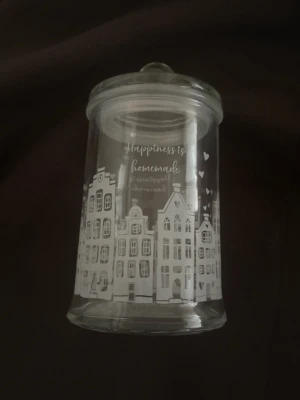 Glasburk från Majas cottage - Säljer en genomskinlig glasburk med lock från Majas Cottage, dekorerad med vita husfasader och texten 'Happiness is homemade'. Perfekt för förvaring av småsaker eller godis. Burken har en rund form och är helt i glas. 15 cm hög