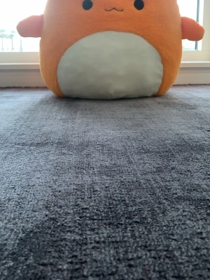 Stor orange Squishmallow plushie - Supermjuk och stor Squishmallow i orange plysch med vit mage och små armar. Den har ett gulligt ansikte broderat framtill och en liten svans baktill. Perfekt som kramkompis eller dekoration i rummet.