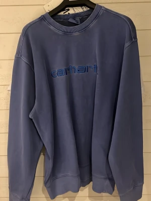 Blå sweatshirt från Carhartt - Säljer en blå sweatshirt från Carhartt med broderad logga framtill i en matchande blå nyans. Tröjan har rund halsringning, ribbade muddar och avslappnad passform. Perfekt för en chill och stilren look.