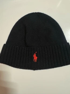Svart mössa från Polo Ralph Lauren - Svart  mössa från Polo Ralph Lauren med klassisk röd broderad logga framtill. Mjuk och skön passform, perfekt för kyliga dagar. Enkel och stilren design som funkar till de flesta outfits. Den är använd 2 gånger och den har blivit lite knottrig.