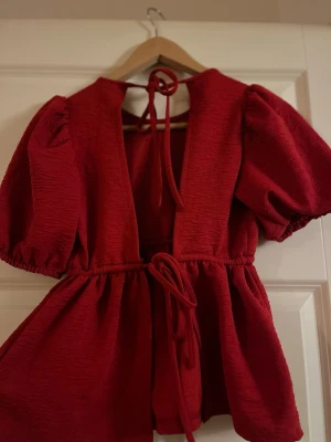 Röd puffärmad blus med knytband - Trendig röd blus med puffärmar och struktur i tyget. Blusen har knytband i midjan och i nacken, samt en djupare urringning fram. Perfekt för dig som vill sticka ut med en färgstark och feminin look.