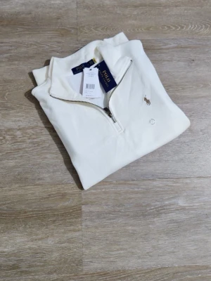 Vit halvzip-tröja från Polo Ralph Lauren - Snygg vit tröja med halv dragkedja från Polo Ralph Lauren. Klassisk design med broderad logga på bröstet och ribbade muddar. Perfekt för en stilren och avslappnad look.