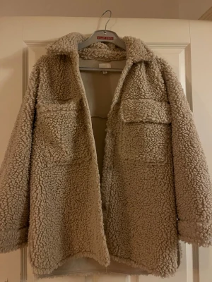 Beige teddyjacka från H&M - Supermysig beige teddyjacka från H&M med två stora bröstfickor och knäppning framtill. Jackan har en oversized passform och är gjord i fluffigt teddy-material som håller dig varm. Perfekt för lager-på-lager och en chill streetstyle-look.