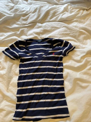 Randig t-shirt från Ralph Lauren - Snygg marinblå och vit randig t-shirt från Ralph Lauren med v-ringning och korta ärmar. Klassisk liten röd logga broderad på bröstet. Tillverkad i mjuk bomull som känns skön mot huden. Skicka gärna prisförslag🤩