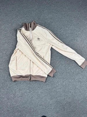 Beige Adidas track jacket - Snygg beige track jacket från Adidas med klassiska bruna ränder längs ärmarna och Adidas-logga på bröstet. Jackan har dragkedja framtill, ribbade muddar och krage samt två fickor med dragkedja. Perfekt för en sportig och avslappnad stil.