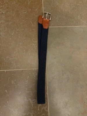 Flätat marinblått bälte Jack & Jones - Snyggt flätat bälte från Jack & Jones i marinblå textil med detaljer i brunt läder och ett klassiskt silverfärgat spänne. Perfekt för att ge din outfit en stilren touch. Passar till både jeans och chinos.