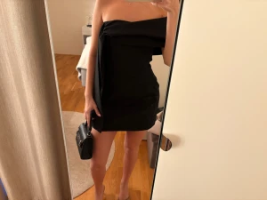 Svart axelbandslös kort klänning - Säljer en svart axelbandslös kort klänning med asymmetrisk volangdetalj framtill. Klänningen har en stilren och modern design som ger en snygg siluett. Perfekt för dig som gillar minimalistisk stil och vill sticka ut. Klänningen är i kostymmaterial och köpt på Zara, prislappen är kvar🥰