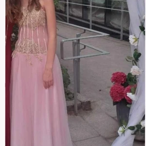 Rosa axelbandslös balklänning med gulddetaljer - Elegant rosa klänning med guldfärgade broderier och pärlor på överdelen. Den är även vadderad så man behöver ingen Bh.Klänningen har en lång, flödande kjol och en vacker, figurnära passform. Perfekt för en glamorös kväll. Jag på första bilden och resterande bilder är lånade, den satt som en M först man jag har sytt om den till en S. Passar dig som är från 160-175. Jag har använt den 1 gång och säljer den eftersom den inte kommer till användning. Köpte den för 1500kr🩷