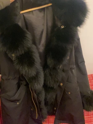 Svart parkas med fuskpälsdetaljer - Stilren svart parkajacka med stora fuskpälsdetaljer runt huva och krage. Jackan har dragkedja, knappar och praktiska fickor framtill. Perfekt för kyliga dagar när du vill hålla dig varm och snygg.