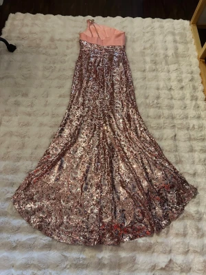 Rosa glittrig långklänning - Rosa, enaxlad långklänning med paljetter och en dekorativ detalj på axeln. Klänningen har en dragkedja baktill och utsvängd kjol som fäller ut i ett lätt släp baktill. Några av de stenar som är på axeln sitter löst men man kan enkelt sy de fast.    Klänningen är kemtvättad men trots det så finns pyttesmå fläckar inifrån se bild som påverkar inte alls utseendet på klänningen.