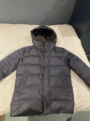 Mörkblå vinterjacka från J Lindeberg - Snygg mörkblå vinter från J Lindeberg med huva och dragkedja. Jackan har en quiltad design och är fodrad för extra värme. Perfekt för kalla dagar och har en stilren look som passar till det mesta. Jackan är i mycket bra skick. Hör av er vid frågor och funderingar!