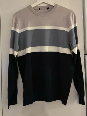 Randig långärmad tröja  - Snygg långärmad tröja med breda horisontella ränder i beige, gråblå, vitt och svart. Tröjan har rund halsringning och är gjord i ett mjukt, tunt material som känns skönt mot huden. Perfekt för en stilren och avslappnad look.