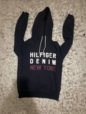 Mörkblå Hilfiger Denim hoodie - Mörkblå hoodie från Hilfiger Denim med stor vit och vinröd text på bröstet. Huva med vita snören. Storlek S, mycket bra skick. Hör gärna av dig!