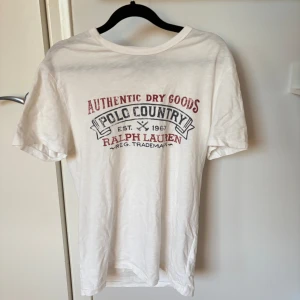 Ralph Lauren t-shirt med tryck - Vit t-shirt från Ralph Lauren med rött och svart tryck på bröstet där det står 'Authentic Dry Goods Polo Country'. Vädligt sällsynt tröja och fint skick! Hör gärna av vid fler funderingar!
